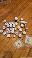 Overview of multiple small jars with white lids containing various coloured powders and gels arranged on a wooden floor.