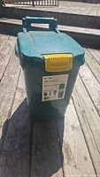 Closed green plastic outdoor storage bin with yellow latch and handle, label visible.