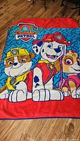 Full view of Paw Patrol fleece blanket showing characters and logo on wooden floor surface.