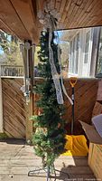 Full view of pre-lit 7ft NOMA pencil Christmas tree with warm white LED lights and decorative bow on top in a wooden porch setting.