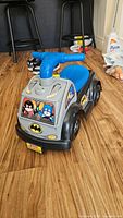Front and side view showing blue handlebar, Batman logos, cartoon character decals, and black wheels with gray rims on plastic ride-on toy.