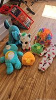 Set of seven plush stuffed animals displayed on wooden floor including Carebear, unicorn, elephant, reversible frog, rainbow animal, orange round plush, and patterned plush.
