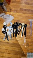 Set of assorted plastic kitchen utensils laid out on a wooden floor, showing various spoons, forks, spatulas, ladles and salad servers in multiple colors.
