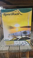 Front of the SportRack Kayak Carrier box showing product name and image of kayak on roof rack at sunset.