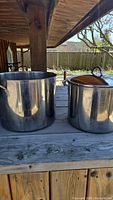Two stainless steel stock pots on wooden table outside, one large without lid, one medium with lid, showing exterior condition and handles.