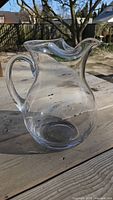 Clear glass pitcher with a curved handle and wide spout shown on wooden table outdoors