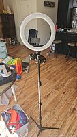Ring light mounted on adjustable black tripod stand in a living room setting, visible phone holder in the center of the ring.