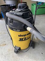 Front side view of the yellow and black Shop-Vac vacuum showing the 10 gallon capacity labeling and attached gray hose coiled around the base with power cord.