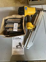 Top-down image of the yellow air framing nailer with attached nail magazine, alongside a box of framing nails and the owner's manual for the tool.