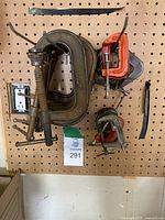 Wide view of various large to small C clamps hanging on a pegboard, showing different sizes and colors including rusted metal and orange painted clamps.