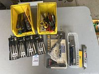 Photo showing two yellow bins containing putty knives with plastic handles, several sets of hobby knives in packaging, Schrade multi-tool, metal utility knife, and a white bin with other utility knives.