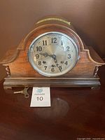 Overall frontal view of the Black Forest Company mantle clock on a dark surface showing the wooden case and metal dial, including winding key in front.