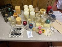 Various candles displayed on a table including pillar candles in beige, cream, and white colors, rose-shaped candles, a red ball candle, tealights, and a snowman figurine candle.