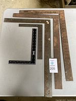 Five carpenter squares of different sizes laid on a table. The squares vary from small black steel to large rusted steel with measurement markings.