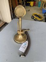 Front view of vintage brass-tone CB base station microphone showing round mesh head, weighted base, and coiled cord on gray surface.