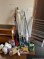 Full view of assorted cleaning implements including mops, brooms, dusters, and a white metal drying rack.