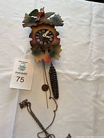 Full view of the cuckoo clock laid flat showing the carved bird and leaf details, Roman numeral face, metal chain, and pine cone weight.