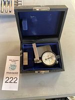 Dial depth indicator in open case showing dial face, adjustable arm, and metal block accessory on blue velvet interior.