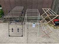 Three drying racks side by side on concrete ground outside next to a fence: large foldable metal with blue joints, a medium white metal drying rack, and a small wooden expandable drying rack.