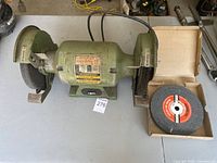 Top-down view of the electric bench grinder on table next to new boxed grinding wheel.