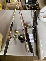 Three fishing rods with reels and an aluminum baseball bat arranged on table.