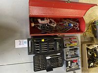 Top view of red metal toolbox filled with copper tubing flaring and bending tools alongside two open plastic tool cases containing various screwdriver bits, drill bits, and hand tools