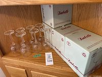 23 clear crystal wine glasses on a wooden surface with 16 glasses in original boxes labeled Isabelle and 7 glasses out of boxes.