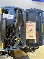 Black Mastercraft reciprocating saw inside molded blue carrying case with yellow latches. Cord is coiled beside the saw and instruction manual is inside the case.