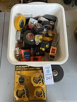 Top view of white bin filled with numerous loose tape measures in various sizes and colors alongside a packaged WorkForce 4-piece tape measure set on a dolly.