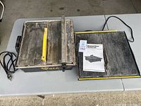 Two used tile saws on a table, one with a yellow safety guard and an instruction manual titled 'Diamond Wheel Cutting Saw'.