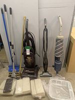 Full view showing three vacuums, three mops, sponges, dish drying rack, and curtain rod against wall.