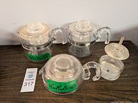 Three Pyrex Flameware glass coffee percolators of different sizes, each with a clear glass lid and large handle, two have stainless steel bases, one is all glass with a separate glass basket accessory.