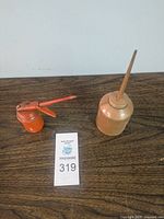 Two vintage oil cans on a wooden surface, one orange with thumb pump, one beige with long spout