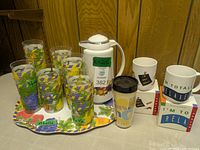 Overview of all items in the lot: 8 acrylic fruit cups with colorful fruit designs, matching rectangular tray with same fruit motif, white thermos with similar fruit pattern and white lid, two white ceramic mugs with text designs, one Zellers travel mug with colored text and black lid.