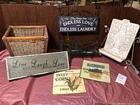 Photo of assorted home decor items arranged on a dark surface including wicker basket, metal and wooden signs, white decorative stand, and metal rack.