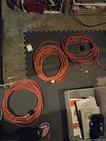 Photo shows three coiled orange electrical extension cords placed on a black and gray foam floor mat and concrete floor, with various objects around them.
