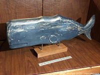 Front side view of the blue painted wooden sperm whale sculpture showing carved eye, mouth, and mounted on a wooden base with metal supports. Ruler for scale.