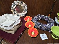 Photo showing collection of summer-themed decorative items including two shell-frame mirrors, orange flower-shaped bowls, beige canvas storage basket, and purple mussel shell wreath on a dark surface.