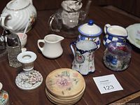 Assorted tea pots, creamers, coasters, candle holder, and glass items arranged on a table, showcasing floral and pastel designs.