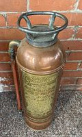 Full standing view of antique copper and brass Guardian fire extinguisher with visible brass label and top handle, photographed against brick background.