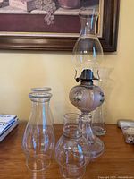 Photo showing two hand-painted blown glass oil lamps with chimneys and a clear glass vase, all positioned in front of a framed painting.