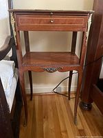 Front view of antique wood side table showing drawer with two brass knobs, pull-out shelf, lower shelf, and curved legs with brass accents.