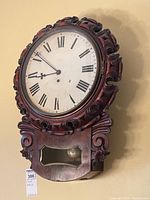 Full view of carved mahogany antique wall clock mounted on wall, showing round dial with Roman numerals, glass-fronted pendulum compartment and decorative wood carvings.