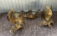 Two brass rooster statues and decorative brass pot arranged on carpet with wall background.