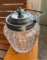 Angled view of the crystal biscuit barrel showing the silver plate lid with decorative engraving and metal handle attached on sides.