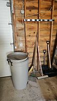 Wide view of the garbage bin and garden tools arranged against a wooden wall in a garage