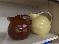 Brown Hall Pottery jug and cream-colored Rouge Mills Pottery jug on shelf, showing overall shape and size.