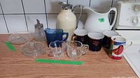 Vintage kitchen glassware and mugs displayed on countertop including footed creamer, sugar bowl, juicer, cobalt blue creamer and mugs, and a ceramic pitcher