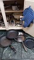 Image showing various frying pans including Rock and Lagostina, ceramic non-stick pan, and parts of other cookware on the floor and inside lower cabinet.