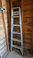 Full view of the 6ft aluminum step ladder standing folded against a wooden wall in a garage.
